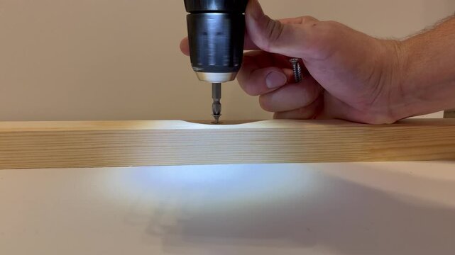 a person's hands using a cordless power drill to drive a metal screw into a light-colored wooden board.
