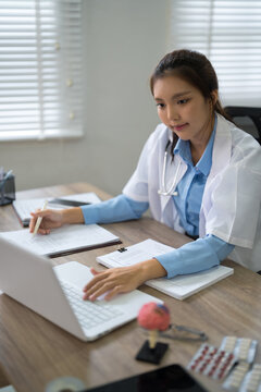 Young Asian female doctor typing on laptop while reviewing patient records at clinic desk with copy space