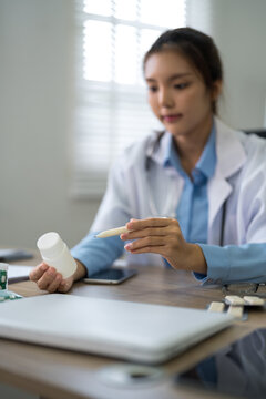 Young Asian female doctor examining prescription medication bottle at clinic desk with natural window light