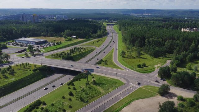 An aerial view shows a complex highway intersection and surrounding roads with driving cars in Vilnius, Lithuania, bordered by dense pine forests.
