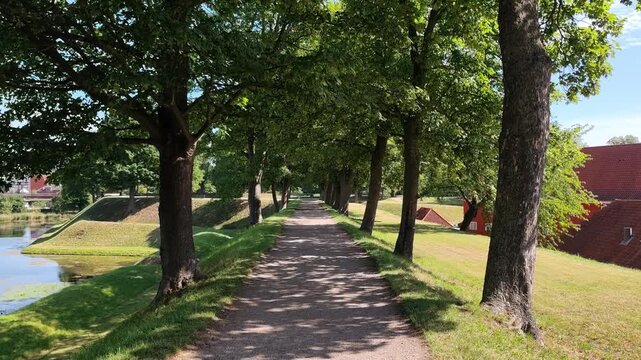 View of the shaded rampart path at Kastellet in Copenhagen, Denmark, lined with leafy trees and overlooking historic red-roofed barracks and a tranquil moat under a clear summer sky.