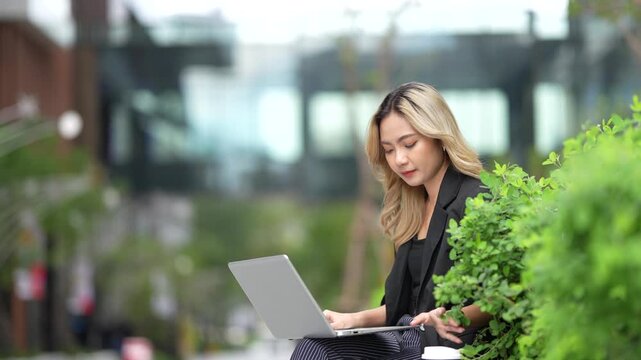 Businesswoman seated behind green shrubs sips coffee then sets down the cup and begins typing on her laptop, framed through leafy foliage in a relaxed outdoor workspace.