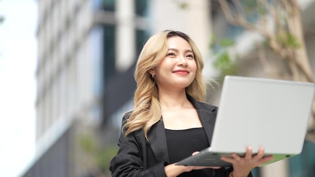 Businesswoman stands holding an open laptop with both hands, reading the screen intently before breaking into a warm smile as the static camera frames her against the city.