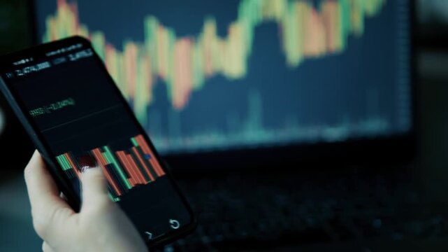 Trader Analyzing Stock Market Candlestick Chart On Smartphone Screen In Front Of Laptop Display