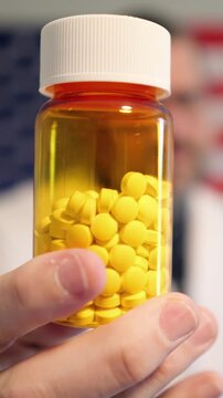 Pharmacist in lab coat holding prescription pill bottle, cash, and crypto against an American flag, symbolizing the influence of money on US medicine