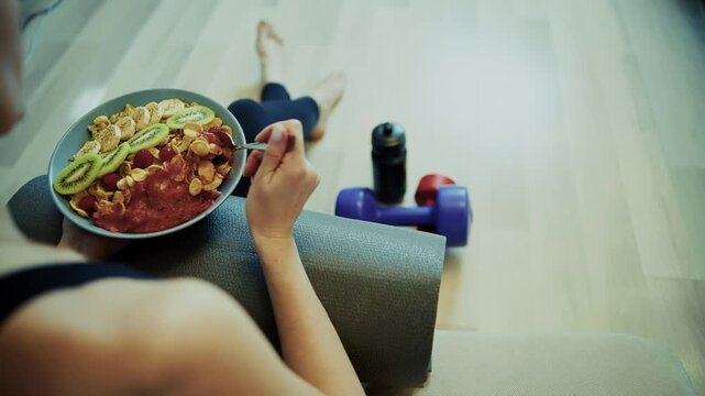 Athlete Eating Fruit Oatmeal Bowl Sitting Near Fitness Mat And Dumbbell 