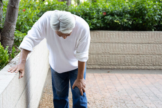 asian senior man with white hair holding painful knee from osteoarthritis, knee joint pain outdoor park scene