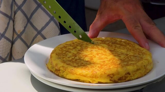 Close-up shot of hands slicing a freshly cooked traditional Spanish potato omelette tortilla de patatas with a knife on a white plate during an outdoor meal.