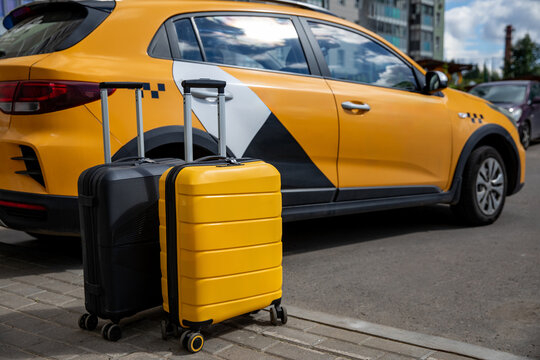Two suitcases, one black and one yellow, positioned beside a yellow taxi car parked on a cobblestone street under a clear blue sky