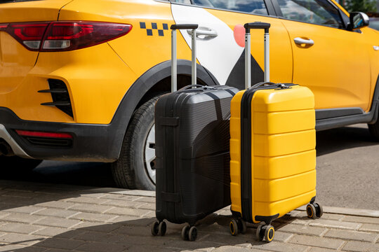 Two suitcases, one black and one yellow, positioned beside a yellow taxi car parked on a cobblestone street under a clear blue sky