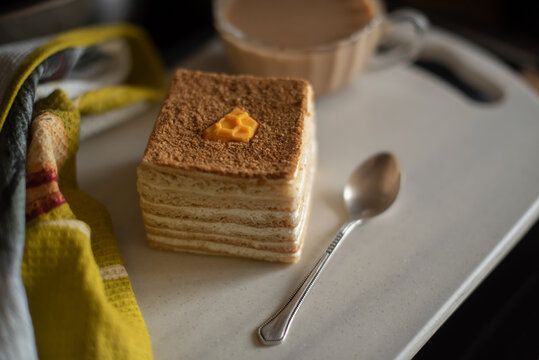 A square honey cake with a honeycomb decoration sits on a white plate. A silver spoon and a bowl are nearby. The scene has ample copy space.