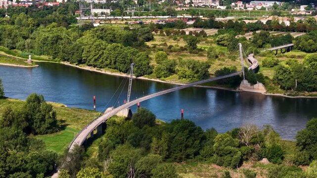 Drone view of Herrenkrug pedestrian bridge crossing Elbe River in Magdeburg, surrounded by green parkland and urban scenery. Summer cityscape shows waterfront, trees, architecture and transport links
