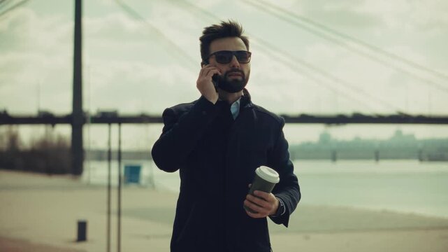 Businessman Talking On Smartphone Holding Coffee Cup. Businessman Walking Near City Bridge