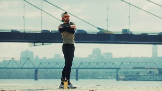 Runner Stretching Arms Warm Up Exercise Near River Bridge In City. Fitness Workout Outdoor