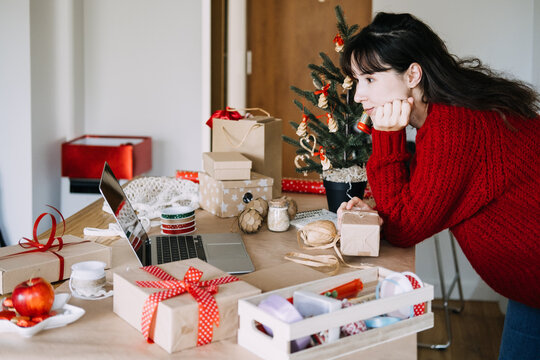 Woman leans on table with laptop, wrapped gifts and small tree while holding parcel. Cozy holiday planning desk, christmas prep workspace, festive desk with laptop notebook