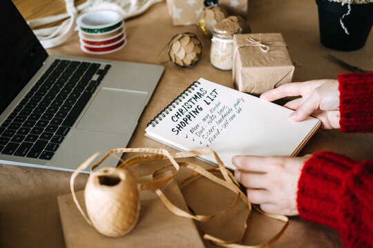 Hands hold notebook with CHRISTMAS SHOPPING LIST beside laptop, wrapped gift and ribbons. Christmas shopping list handwritten, gift list for family, holiday shopping checklist laptop