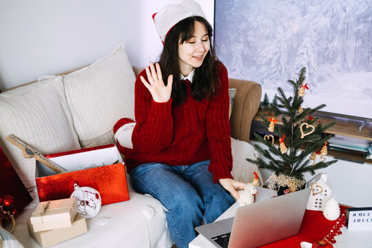Woman in Santa hat waves at laptop on sofa beside decorated tree and HOLLY JOLLY sign. Christmas video call laptop, holiday virtual celebration, waving at video call christmas