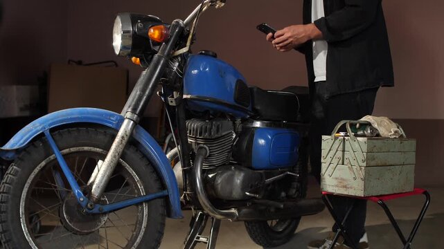 Young person checking repair instructions on a smartphone while working on a vintage blue motorcycle with a toolbox, standing in a garage, focusing on retro vehicle maintenance