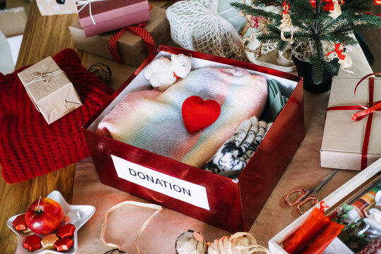 DONATION box filled with clothes stands on table beside Christmas tree and wrapped gifts. Christmas donation box, holiday charity clothing drive, donating warm clothes winter