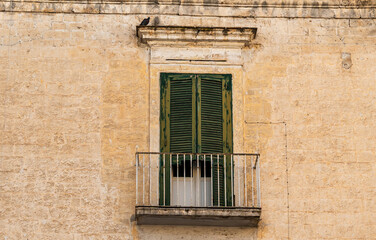 Fototapeta premium Old stone building facade in Bari, Italy with weathered green wooden shutters on balcony door, rustic limestone wall texture and iron railing, nobody
