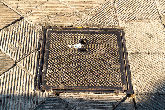Square manhole cover with diamond pattern grid set into paved sidewalk in Bari, Italy, close-up view of urban infrastructure and street surface detail