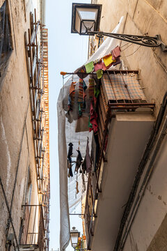 Bari Italy old town narrow alley with colorful laundry hanging between aged building facades, balcony with reed blind and street lamp overhead, Mediterranean daily life