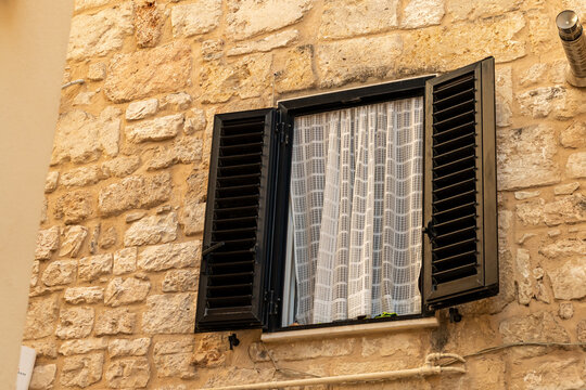 Traditional stone wall building with open black shutters and white lace curtain in Bari, Italy, Mediterranean architecture detail, concept of historic European residential facade