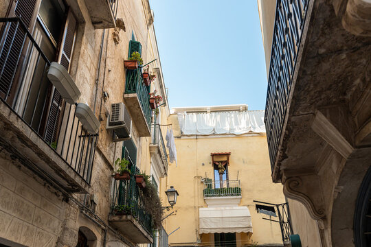 Bari Italy old town narrow street with aged residential buildings featuring balconies, potted plants and hanging laundry under clear blue sky, Mediterranean urban scene