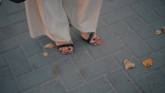 Low downward view of lower legs and feet walking across rectangular stone pavers, with pant hem swaying, sandal heels touching ground, and scattered autumn leaves adding seasonal street style