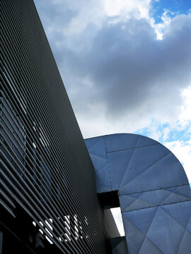 A modern building with a slatted facade and a curved metal section is shown against a cloudy sky.