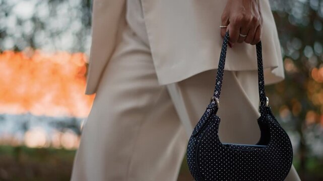 Woman walking outdoors at sunset holding black beaded crescent handbag by handle, with relaxed stride, gentle arm swing, elegant outfit, warm glowing water, and soft evening light, with handbag, bag