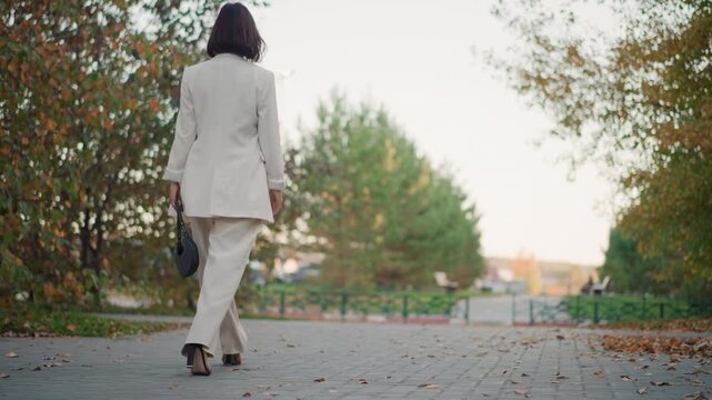 Rear view of woman walking away along tree lined paved park path wearing white tailored suit and carrying handbag, showing steady stride, heel lift, soft daylight, and calm elegant outdoor mood