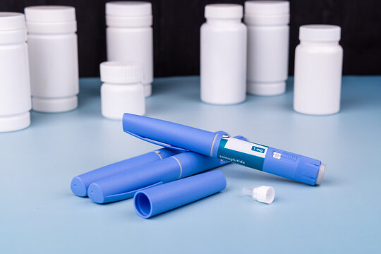 Blue Semaglutide GLP-1 medication pens on a light blue table with white pill bottles background, dark backdrop medical setup