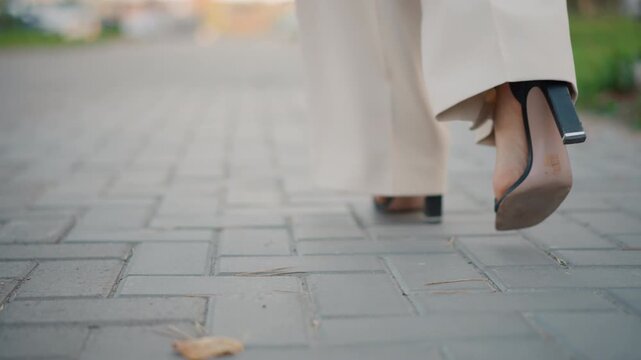 Rear low-angle view shows lower legs and high heels in mid-stride on paved sidewalk, with light skirt hem swaying, heel lift, and steady forward movement in soft daylight, with heel, walking