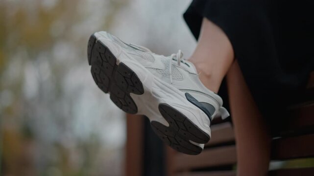 Light-colored sneaker shown from underside while foot rests on wooden park bench with slight toe lift indicating gentle swing or paused motion; rubber tread, textured outsole, and laces are