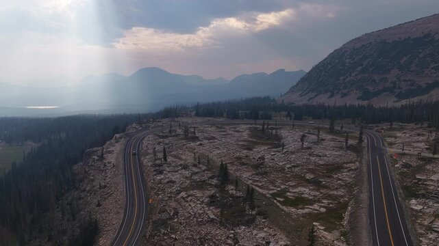 Aerial of curve on highway 150 in Wasatch National Forest in Uinta Mountains winding through an expansive high altitude terrain with dramatic smoky skies in summer