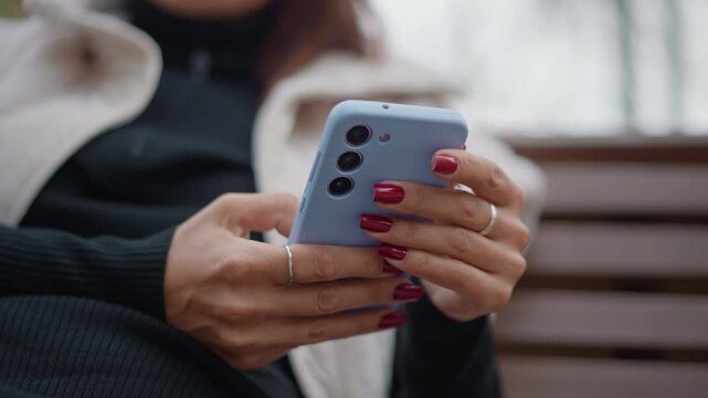 Close view of mature adult hands holding pale blue smartphone on wooden park bench, thumbs tapping and scrolling with dark red manicure, showing focused casual outdoor mobile use in soft daylight