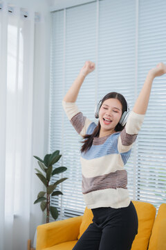Young Woman Dancing to Music: A joyful young woman wearing headphones dances enthusiastically in her living room, her arms raised in celebration as she enjoys her favorite tunes.