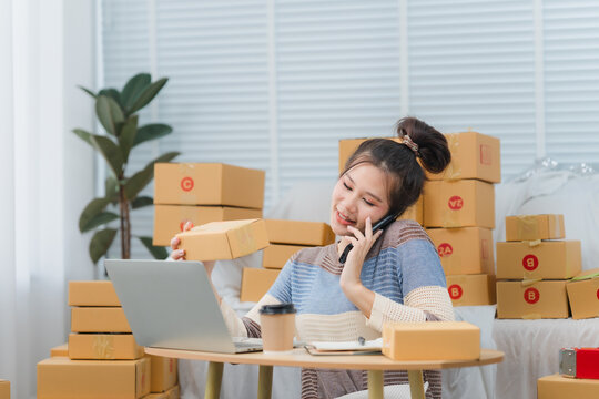E-commerce Hustle: Young Asian entrepreneur managing orders and logistics on phone and tablet, surrounded by shipping boxes in her home office.	
