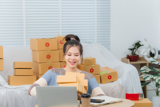 Small Business Owner at Work: A young woman smiles as she prepares packages for shipping, surrounded by stacks of boxes. She is working at her desk with a laptop, a cup of coffee, and order forms.