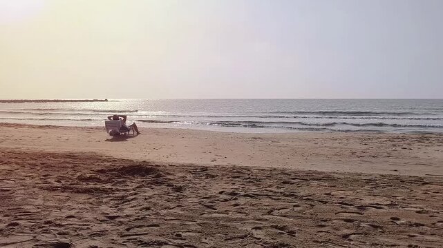 Unrecognizable person sits on a beach chair placed on the sand while looking at the sea as the waves move gently in the background, representing peaceful summer vacations and relaxation