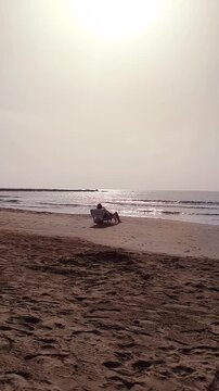 Vertical video of an unrecognizable person sitting on a beach chair on the sand while facing the sea, with gentle waves moving in the background, representing summer holidays and relaxation
