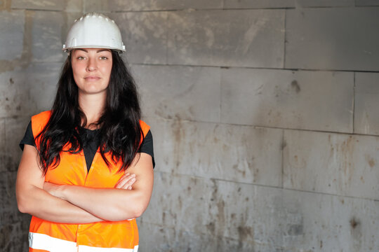 Young woman in builders hi vis vest and helmet posing next to bare bricks wall - females in construction concept