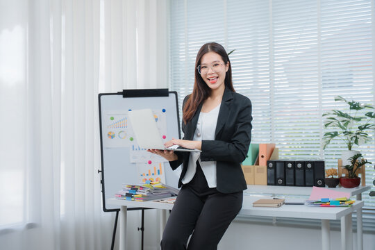 Confident Businesswoman Presenting: A professional businesswoman in a sharp suit stands confidently beside a whiteboard, laptop in hand.