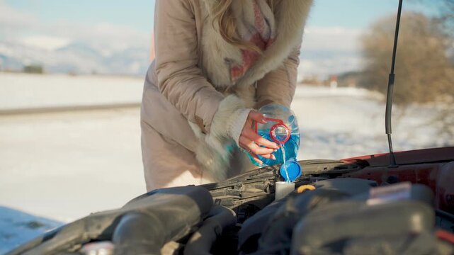 Woman pouring anti freeze windshield fluid into car on cold winter day, snow covered fields and mountains blurred background, closeup to hands holding bottle with blue liquid