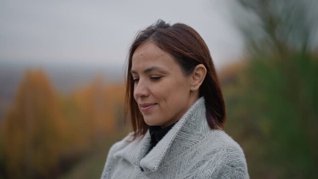 Woman wrapped in chunky knitted shawl stands chest-up on muted autumn hillside, head turned slightly right with composed expression and faint smile, conveying calm confidence and quiet seasonal mood