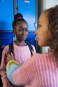 Two teen females in pink sweaters standing at blue lockers one crying while friend holding backpack
