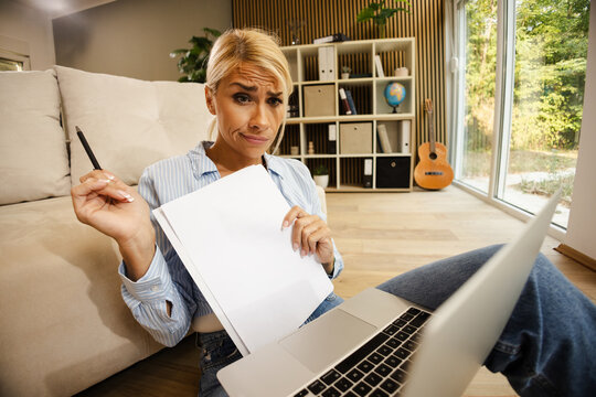 Woman Working on a Laptop in a Small Office Space While Holding a Pen and Blank Paper in a Business Setting