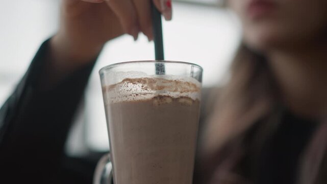 Tall glass of chocolate milkshake or iced coffee held near rim while person stirs or sips with straw; visible upward motion of straw creates surface ripples, frothy foam, and small bubbles while