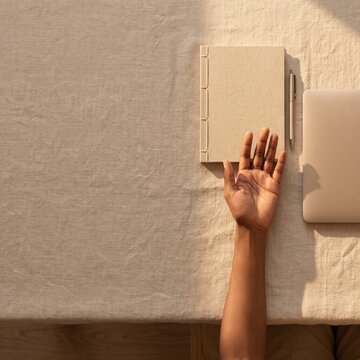 Top view of a person's hand resting on a linen table beside a notebook, pen, and laptop in warm light with copy space